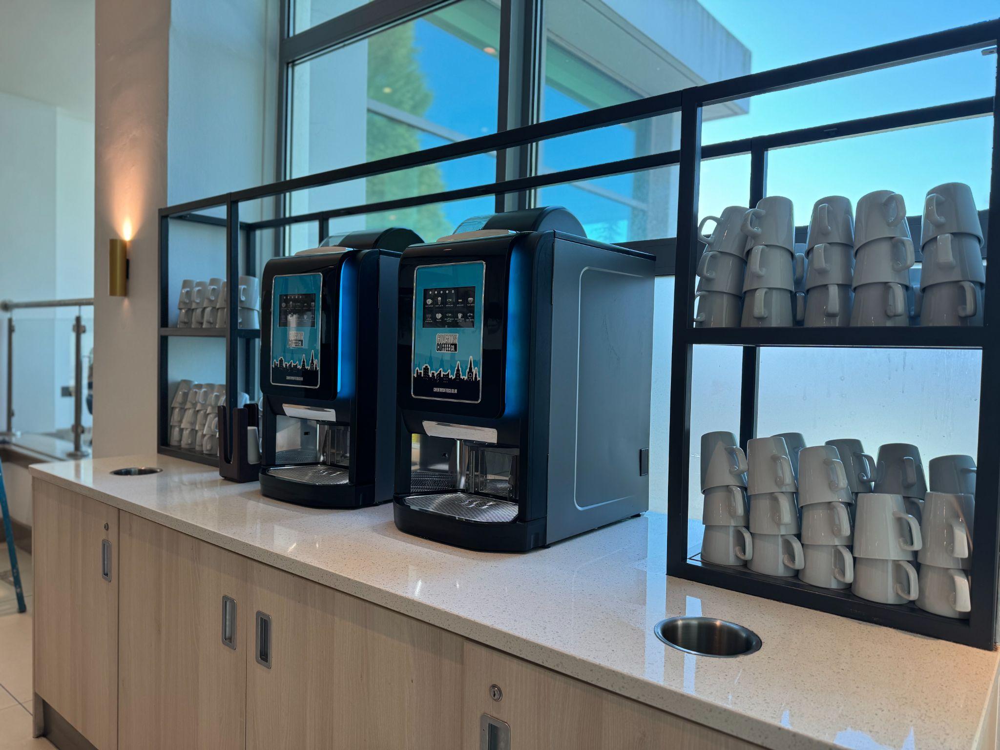 Two Coventry Coffee Co bean-to-cup machines on a hotel buffet counter with stacks of cups behind in a glass shelving unit