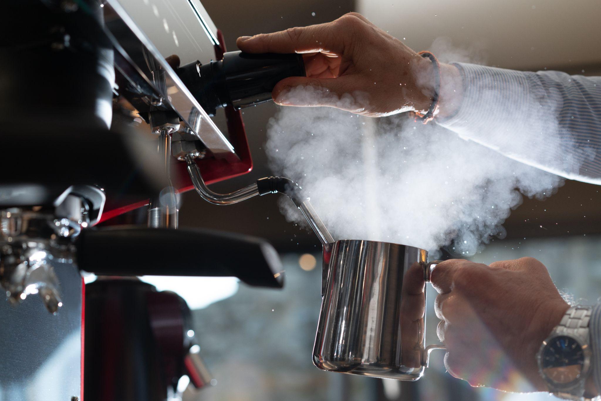Barista steaming milk on a commercial Gaggia espresso machine, fresh steam rising from a stainless steel jug